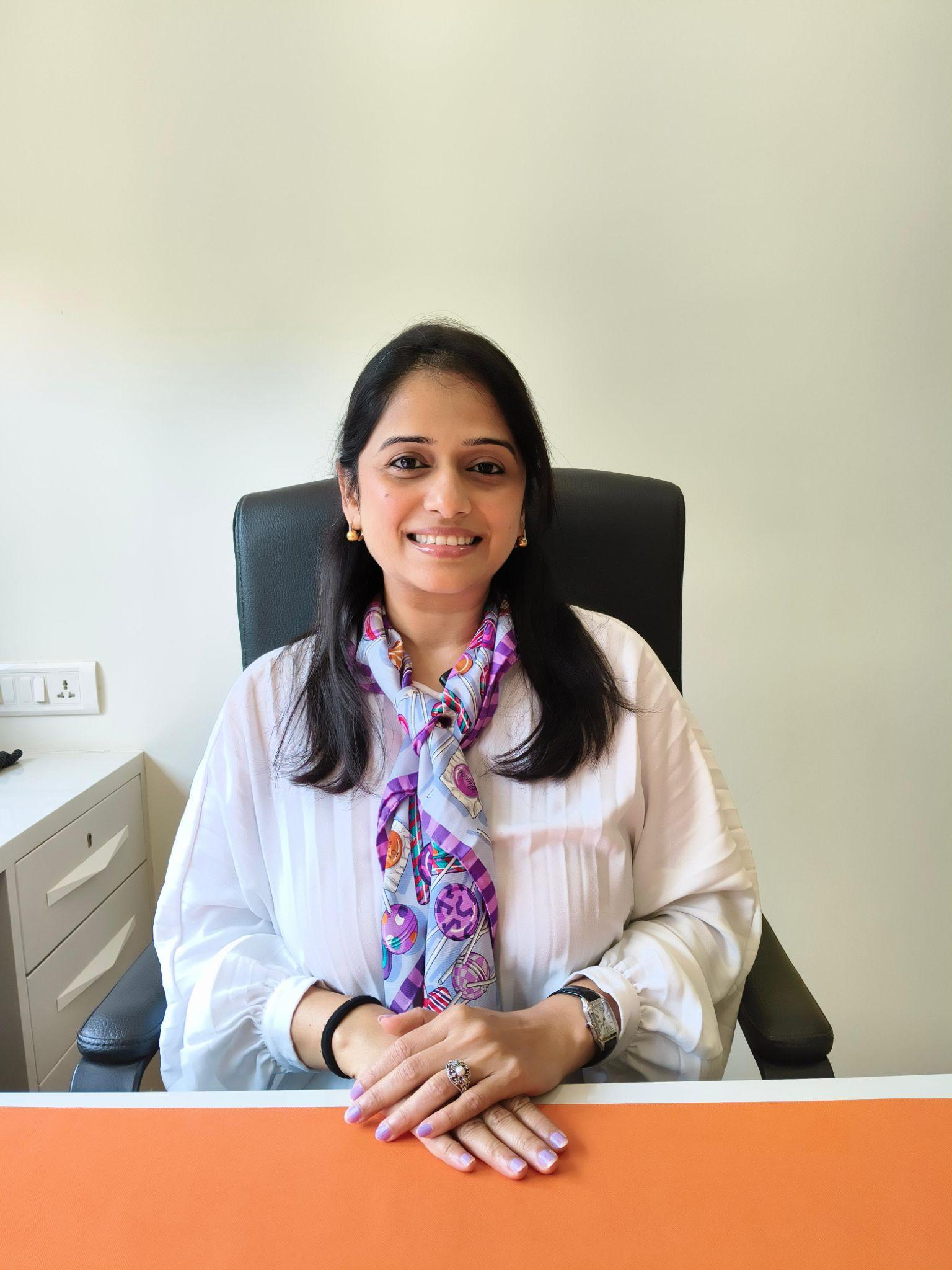 Smiling woman seated in an office chair behind a desk, hands resting on an orange desk mat, wearing a white blouse and colorful scarf.